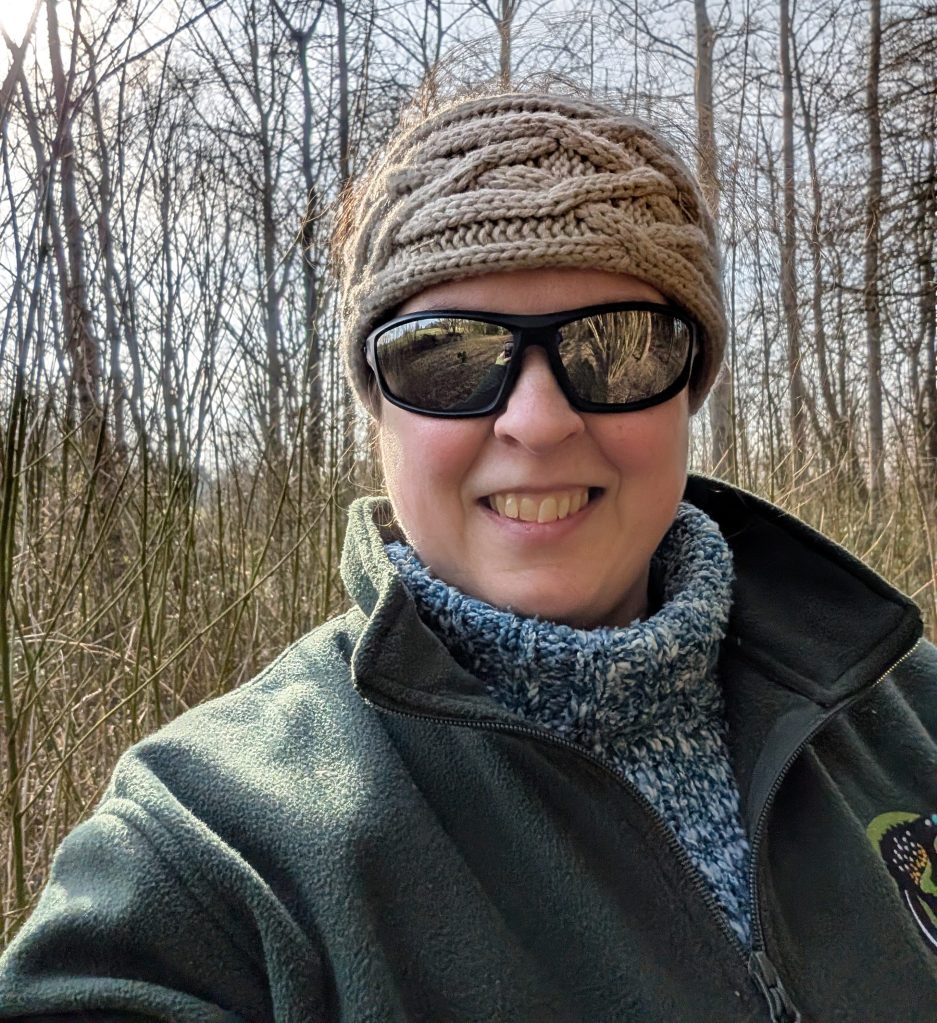 Photograph of Siobhan Brocklehurst outdoors in a woodland, smiling, wearing sunglasses and a woolen headband.